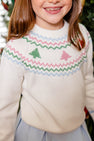 A child smiles in front of a blurred background, dressed in The Beaufort Bonnet Company’s Isabelle’s Fair Isle Sweater—crafted from 100% cotton in the Palmetto Pearl colorway with charming green and pink tree patterns and zigzag designs—paired with a light blue skirt.