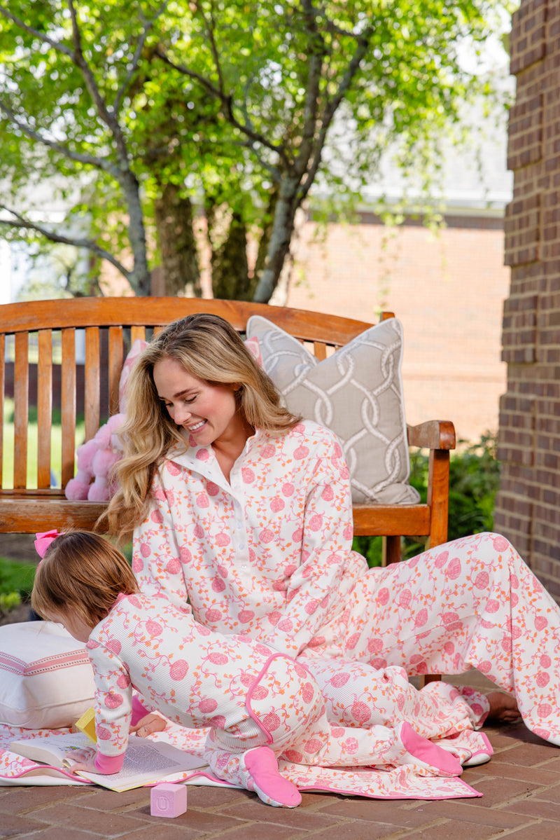 A woman dressed in The Beaufort Bonnet Company’s Noelle’s Night Night - Gourd-geous (Palmetto Pearl) with Palm Beach Pink sits on a wooden bench, smiling at a young child in matching pajamas who is playing on the ground with books and toys. They are outdoors, beside a brick wall and lush green trees.
