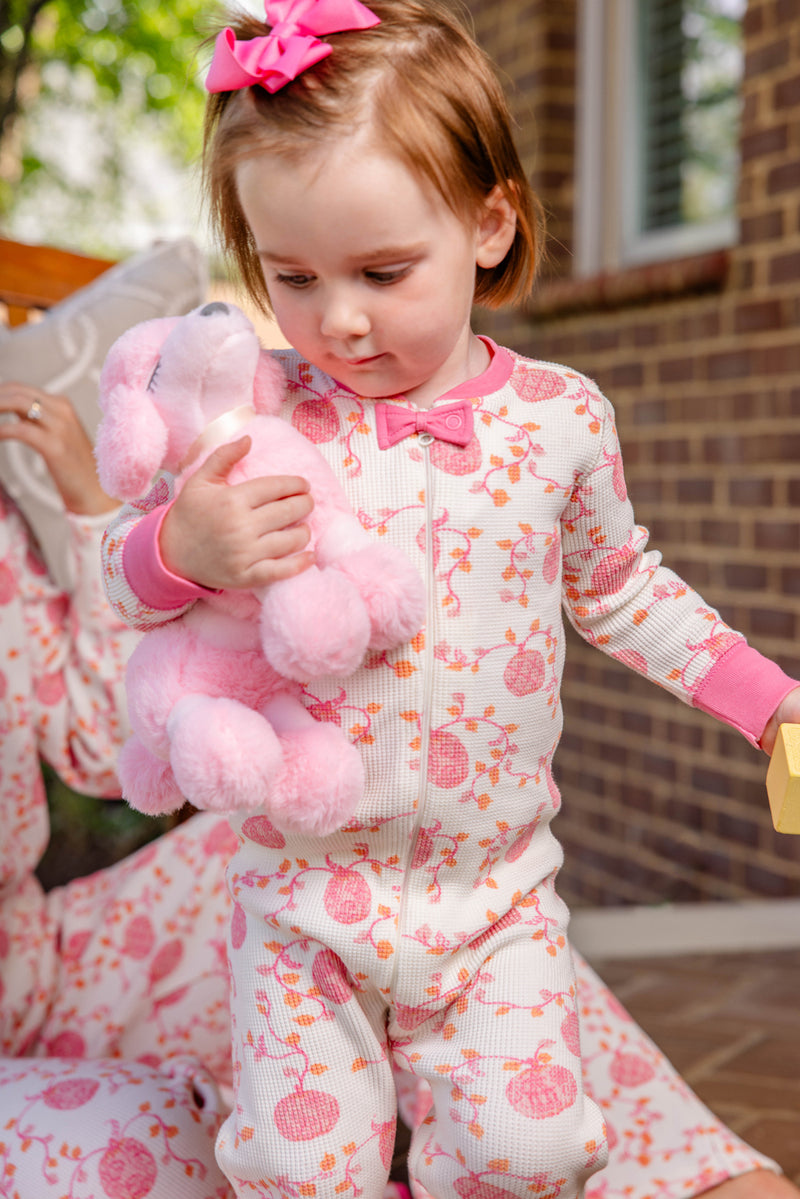 A young girl wearing The Beaufort Bonnet Company’s Noelle’s Night Night - Gourd-geous (Palmetto Pearl) with Palm Beach Pink stands outside by a brick wall. She holds a pink stuffed animal, her hair adorned with a matching pink bow, and looks down thoughtfully in her cozy sleepwear.