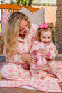 A woman and a young girl in matching pink and white pajamas sit on a porch, both smiling as they relax together on the Silent Night Throw - Gourd-geous (Palmetto Pearl) with Romany Rose by The Beaufort Bonnet Company. The girl holds a pink stuffed animal and wears a pink bow in her hair.