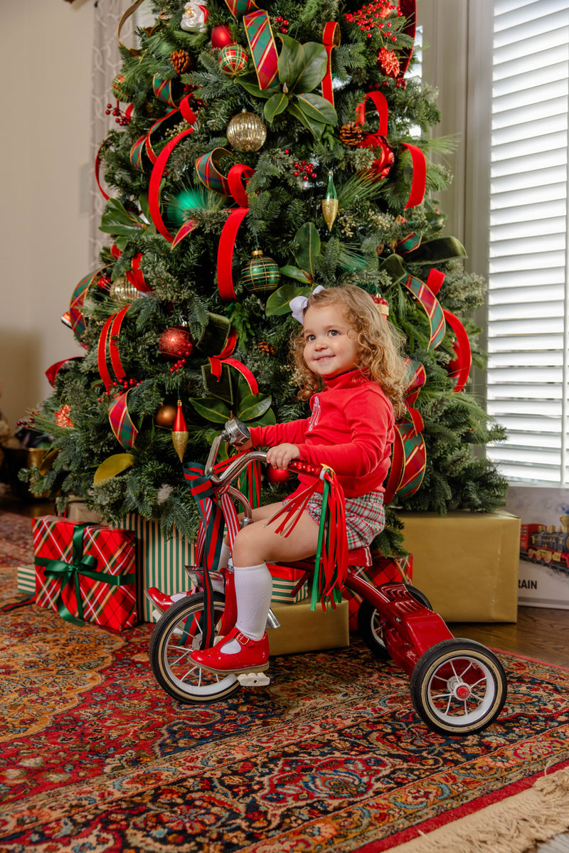 A young child wearing the Natalie Knickers - Wicker Park Plaid with Richmond Red by The Beaufort Bonnet Company sits on a red tricycle in front of a decorated Christmas tree with wrapped gifts underneath.