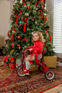 A young child wearing the Natalie Knickers - Wicker Park Plaid with Richmond Red by The Beaufort Bonnet Company sits on a red tricycle in front of a decorated Christmas tree with wrapped gifts underneath.