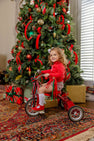 A young child wearing the Natalie Knickers - Wicker Park Plaid with Richmond Red by The Beaufort Bonnet Company sits on a red tricycle in front of a decorated Christmas tree with wrapped gifts underneath.