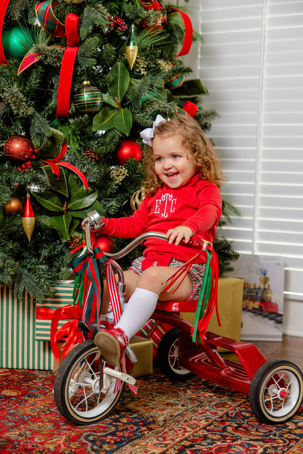 A young girl with curly hair and a white bow rides a red tricycle by a decorated Christmas tree with presents underneath. She wears The Beaufort Bonnet Company’s Mitzy Sue Slacks in Richmond Red, white socks, and smiles happily.