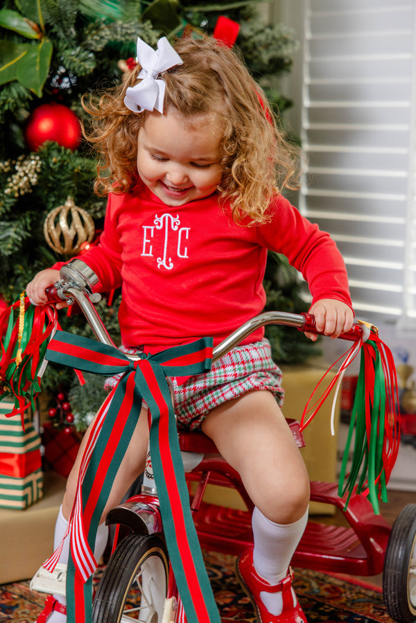 A young child with curly hair and a white bow is dressed in The Beaufort Bonnet Company’s Natalie Knickers in Wicker Park Plaid with Richmond Red, riding a tricycle adorned with red and green ribbons in front of a Christmas tree decorated with ornaments and wrapped gifts.