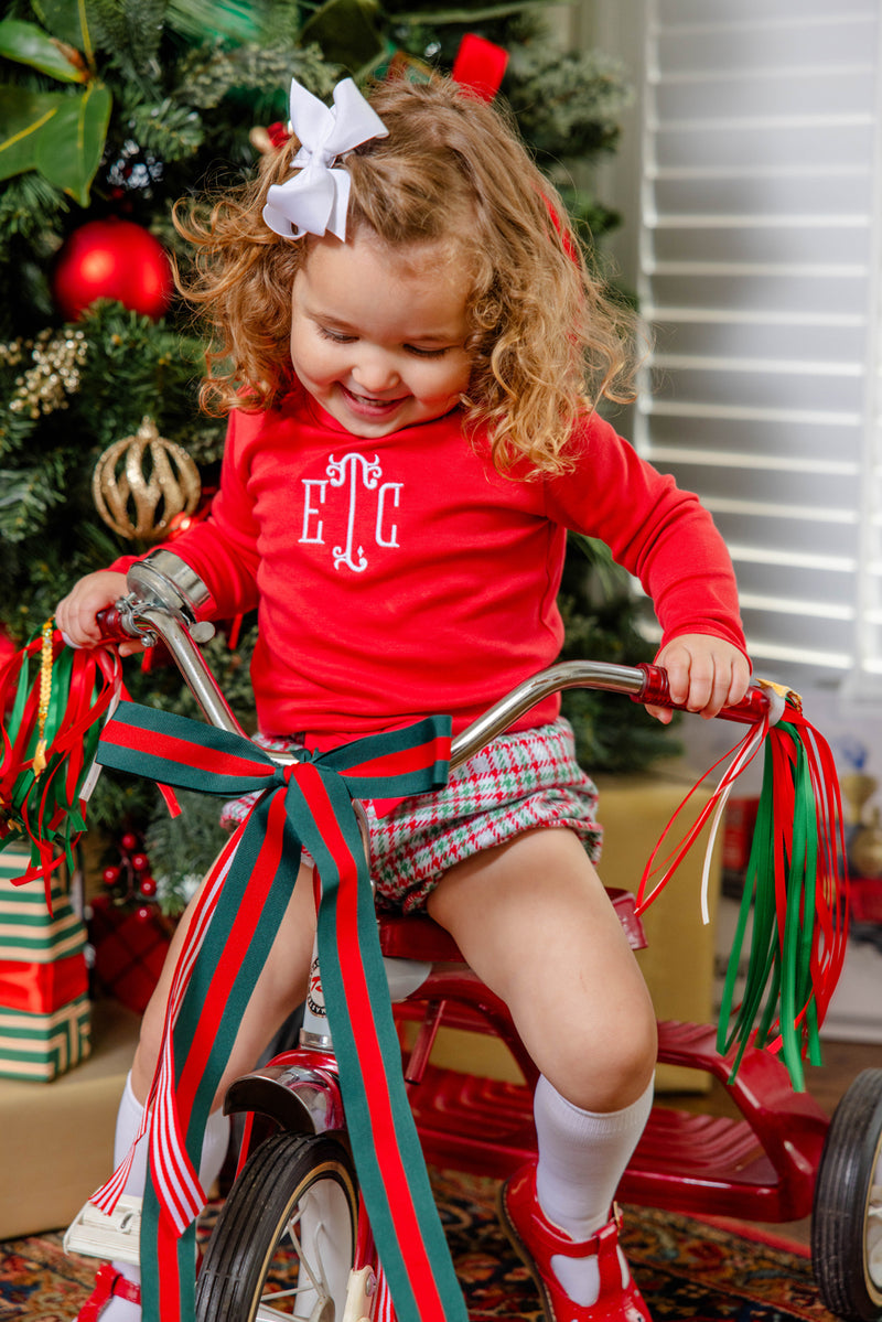 A young child with curly hair and a white bow is dressed in The Beaufort Bonnet Company’s Natalie Knickers in Wicker Park Plaid with Richmond Red, riding a tricycle adorned with red and green ribbons in front of a Christmas tree decorated with ornaments and wrapped gifts.