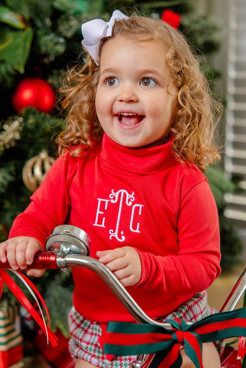 A smiling young girl with curly hair and a white bow sits on a tricycle in front of a decorated Christmas tree, wearing The Beaufort Bonnet Company’s Mitzy Sue Slacks in Richmond Red with white monogram letters and plaid accents.