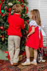 A young girl wearing The Beaufort Bonnet Company’s Mary Dal Dress in Richmond Red & Worth Avenue White with Christmas Tree decorates for the holidays alongside a boy, as ornaments adorn the tree and presents and a toy wagon rest beneath it.