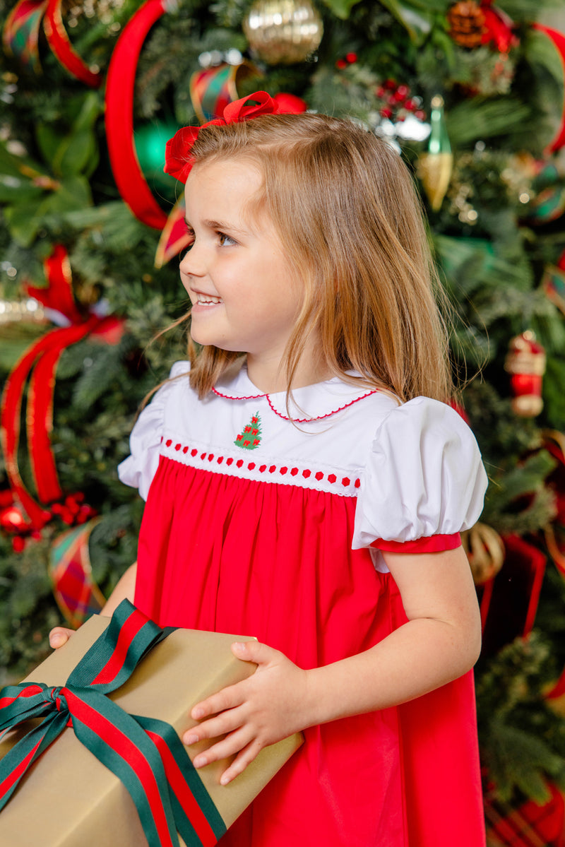 Wearing the Mary Dal Dress in Richmond Red & Worth Avenue White with Christmas Tree from The Beaufort Bonnet Company, a smiling young girl holds a gold gift box with a green and red ribbon, standing in front of a beautifully decorated Christmas tree.