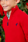 A smiling child wears The Beaufort Bonnet Company’s Hayden Half-Zip Sweater in Richmond Red, featuring a Nantucket Navy stork logo and a classic quarter-zip style, layered over a green plaid shirt. Crafted from 100% cotton, this holiday sweater stands out amid blurred greenery and festive decorations in the background.