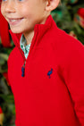 A smiling child wears The Beaufort Bonnet Company’s Hayden Half-Zip Sweater in Richmond Red, featuring a Nantucket Navy stork logo and a classic quarter-zip style, layered over a green plaid shirt. Crafted from 100% cotton, this holiday sweater stands out amid blurred greenery and festive decorations in the background.