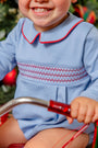 A smiling child in a Long Sleeve Bradford Bubble - Barrington Blue with Richmond Red Smocking by The Beaufort Bonnet Company sits on a tricycle, with a decorated Christmas tree in the background.