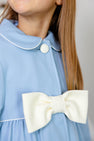 A young girl wearing The Beaufort Bonnet Company’s Pippa Peacoat in Barrington Blue with Palmetto Pearl accents. The holiday coat features a large white bow on the front, as well as a white button and white piping. Only the lower half of her face and her straight brown hair are visible.