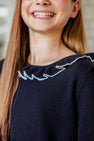A person with long brown hair smiles while wearing The Beaufort Bonnet Company’s Reece Ruffle Sweater in Newport Night with Palmetto Pearl, featuring white zigzag embroidery near the neckline.