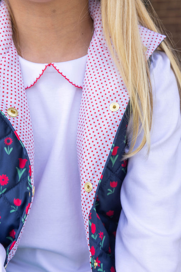 A child wears Emma’s Elbow Patch Top - Worth Avenue White with Richmond Red Flowers, a long-sleeved Pima cotton shirt under a navy blue quilted vest featuring red flower patterns, a red-and-white polka dot lining, and elbow patches.