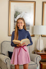 A smiling girl with long hair stands indoors, holding an open book. She wears The Beaufort Bonnet Company’s Tatum’s Turtleneck & Onesie (Unisex) in Nantucket Navy and a pink skirt with a ribbon. Behind her are a framed bird print, a lamp, and a cushioned chair.
