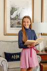 A smiling girl with long brown hair, wearing a navy turtleneck and The Beaufort Bonnet Company’s Parson Pleated Skirt in Begley Berry Tweed, stands holding an open book. She is in a cozy living room with a framed bird painting and a monogrammed pillow on a chair.