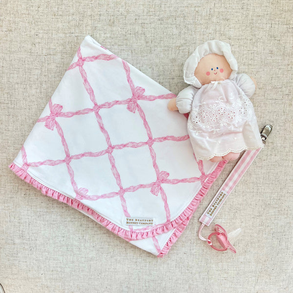 A small stuffed doll wearing a white bonnet and dress, attached to a Parker Paci Clip - Pink Greenbrier Gingham with Palm Beach Pink and a pink baby pacifier.
