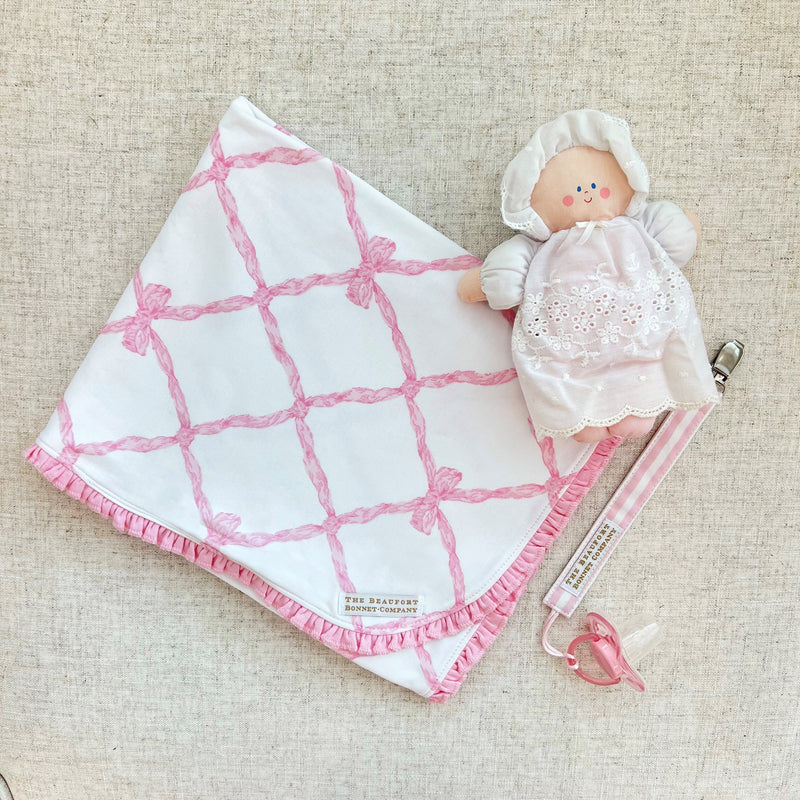 A small stuffed doll wearing a white bonnet and dress, attached to a Parker Paci Clip - Pink Greenbrier Gingham with Palm Beach Pink and a pink baby pacifier.