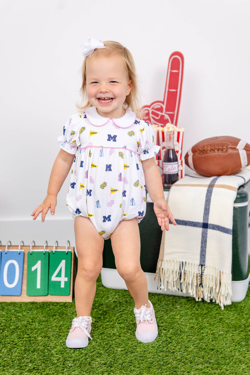 A smiling toddler stands on artificial grass wearing The Beaufort Bonnet Company’s Michigan Britt Bubble - Bold in Blue with Hamptons Hot Pink, crafted from soft Pima Cotton Knit. Behind her are a football, a foam finger, a striped blanket, a drink, and a 2014 sign. She pairs her outfit with pink shoes and a white bow.