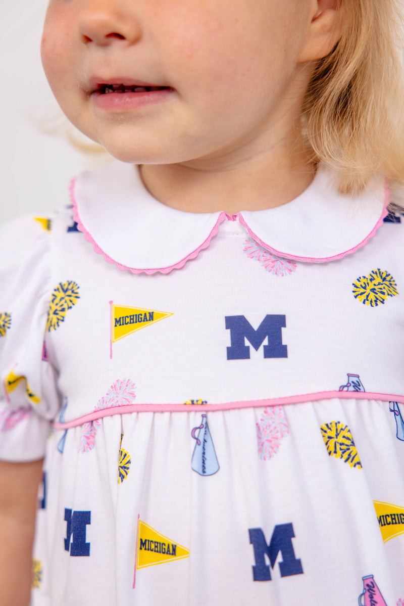 A young child wears The Beaufort Bonnet Company’s Michigan Britt Bubble - Bold in Blue with Hamptons Hot Pink, a soft Pima Cotton Knit dress adorned with blue Ms, yellow Michigan pennants, and blue and yellow pom-poms. The dress features pink trim and a white Peter Pan collar, with only the child’s lower face visible.