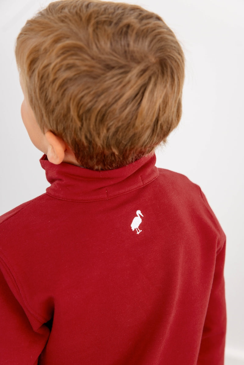 A young child with light brown hair is seen from behind, wearing The Beaufort Bonnet Company’s USC Hayword Half-Zip in South Carolina Garnet with Worth Avenue White, which features a small white bird logo on the upper back near the collar. The plain, light-colored background subtly reflects Gamecock style.