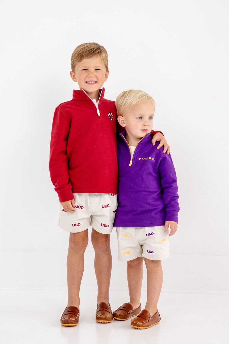 Two young boys stand together against a white background. One is smiling in The Beaufort Bonnet Company’s USC Hayword Half-Zip in South Carolina Burgundy with Worth Avenue White, paired with USC shorts, while the other wears a purple pullover with LSU shorts. Both are barefoot and happy.