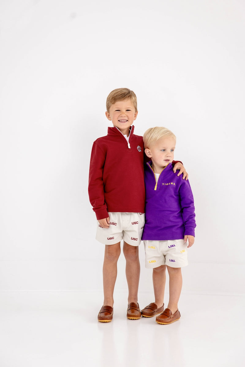 Two young boys stand together, smiling. One wears The Beaufort Bonnet Company’s USC Hayword Half-Zip in South Carolina Garnet with Worth Avenue White, paired with matching shorts in Gamecock style. The other wears a purple pullover and shorts featuring LSU colors. Both have light brown shoes and stand against a plain white background.