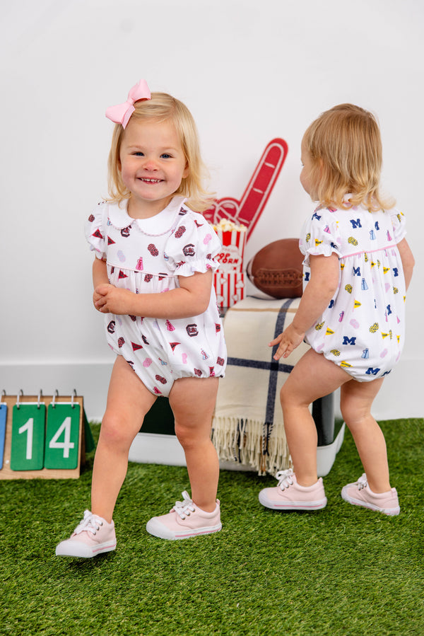 Two toddlers in sports-themed rompers play on green turf. One, dressed in The Beaufort Bonnet Company’s Michigan Britt Bubble - Bold in Blue with Hamptons Hot Pink and wearing a pink bow, smiles at the camera while the other faces away. Props include a football, a foam finger, a calendar with 14, and a blanket.