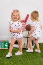 Two toddlers in sports-themed rompers play on green turf. One, dressed in The Beaufort Bonnet Company’s Michigan Britt Bubble - Bold in Blue with Hamptons Hot Pink and wearing a pink bow, smiles at the camera while the other faces away. Props include a football, a foam finger, a calendar with 14, and a blanket.