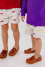 Two children stand side by side on a white floor, one wearing The Beaufort Bonnet Company’s South Carolina Critter Sheffield Shorts in Saratoga Stone and the other in LSU shorts, both sporting brown loafers. Their upper bodies are not visible in the image.
