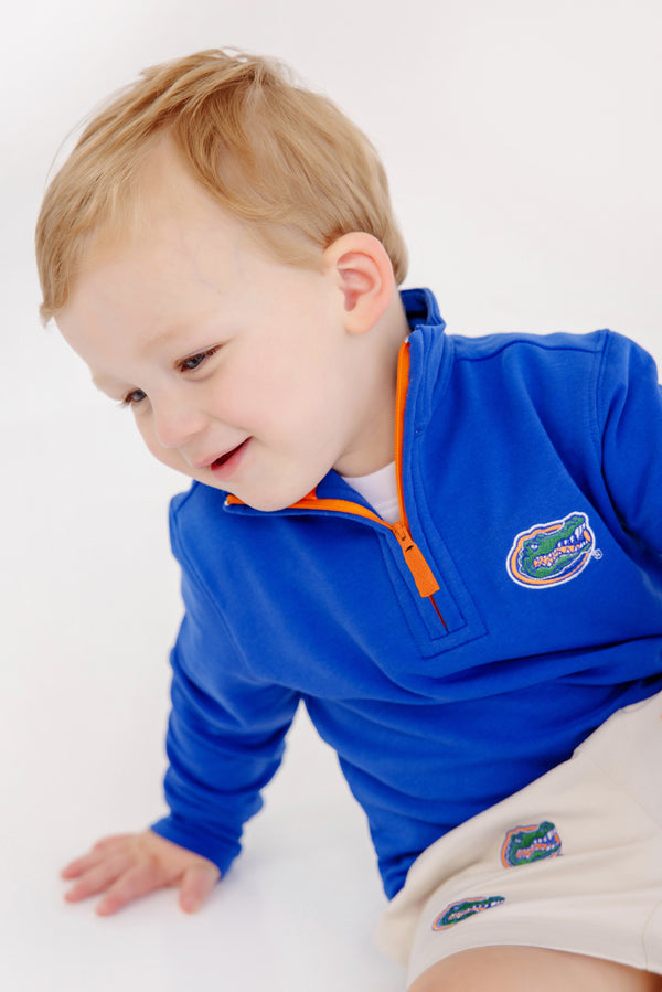 A young child with light hair is smiling while wearing The Beaufort Bonnet Company’s Florida Hayword Half-Zip in vibrant Florida Blue with Florida Green and Orange accents, featuring an orange zipper and the University of Florida Gators logo, paired with matching shorts, against a white background.