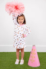 A smiling young girl wearing The Beaufort Bonnet Company’s Auburn Searcy Skort in Tiger Thrill with Worth Avenue White holds a large pink pom-pom above her head, standing beside a pink cheer megaphone labeled TBBBC on vibrant green turf.