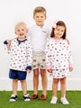 Three young children stand smiling on grass against a white background, dressed in coordinated school spirit outfits. The boy in the middle wears The Beaufort Bonnet Company’s Auburn Prim & Proper Polo in Worth Avenue White, featuring an embroidered Auburn logo, while the others sport patterned sweatshirts with matching shorts and a skirt.