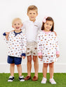 Three young children stand smiling on grass against a white background, dressed in coordinated school spirit outfits. The boy in the middle wears The Beaufort Bonnet Company’s Auburn Prim & Proper Polo in Worth Avenue White, featuring an embroidered Auburn logo, while the others sport patterned sweatshirts with matching shorts and a skirt.