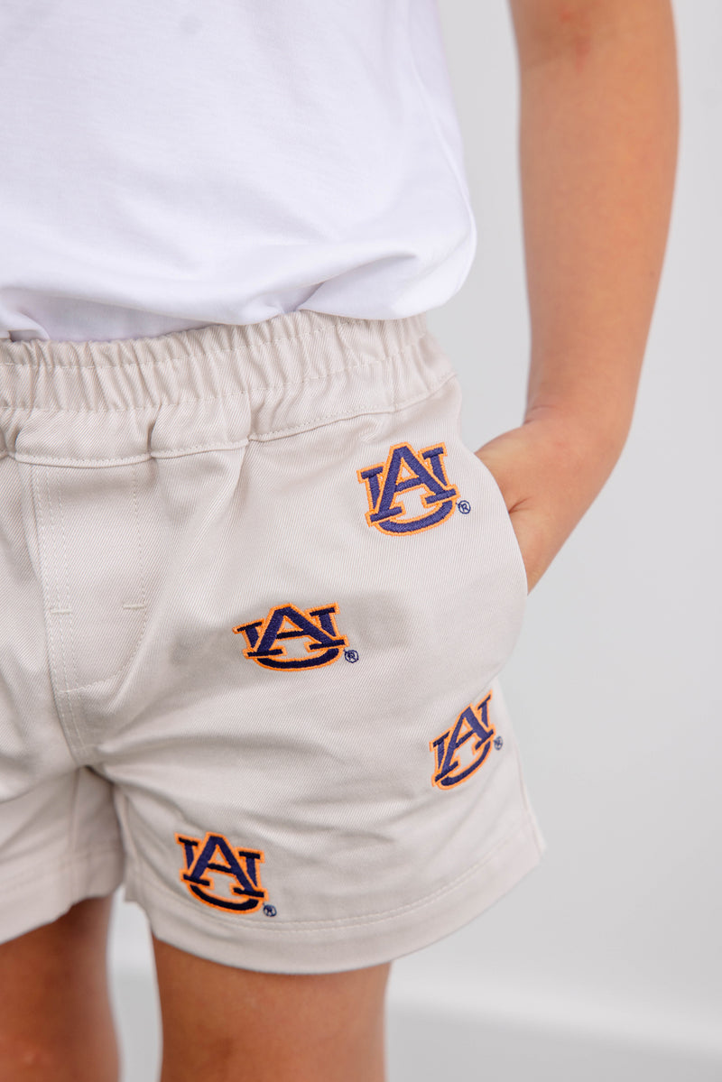 A child wearing The Beaufort Bonnet Company Auburn Critter Sheffield Shorts in Saratoga Stone, adorned with multiple Auburn University logos, stands with one hand in their pocket and a white shirt.
