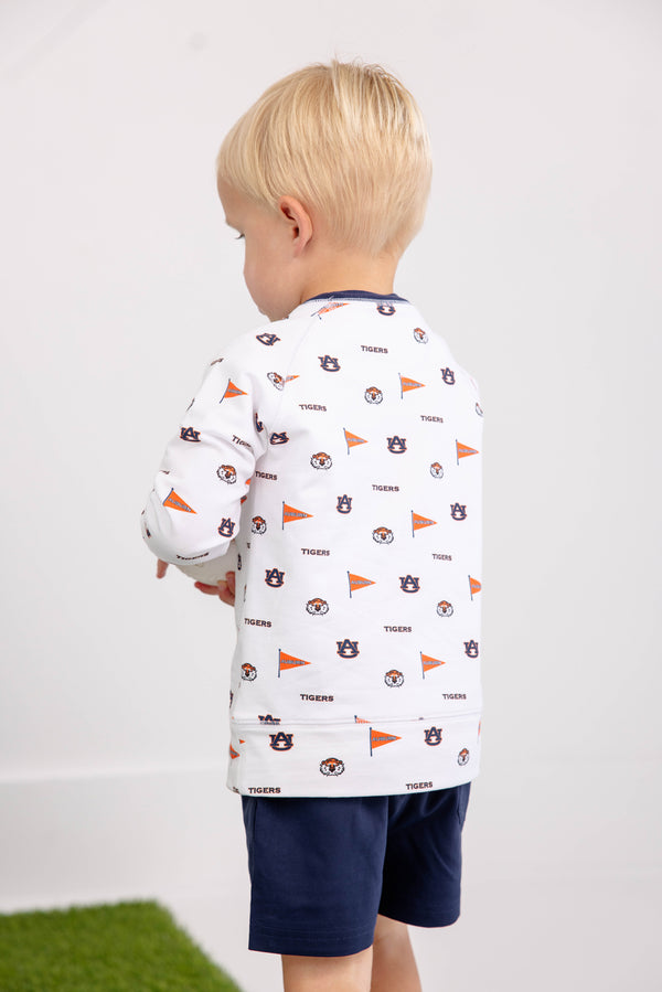 A young child with blonde hair, wearing The Beaufort Bonnet Company’s Auburn Cassidy Comfy Crewneck - Tiger Thrill with Auburn Blue—featuring logo prints, orange flags, and the word TIGERS—stands facing away near a patch of green grass, paired with navy blue shorts.