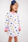 A young girl smiles, dressed in The Beaufort Bonnet Company’s Michigan Searcy Skort - Bold in Blue with Worth Avenue White. Her white outfit features pink cuffs and a playful pattern of blue Ms, pink dresses, yellow flags, and pom-poms. She wears a large navy blue bow in her hair and stands against a plain white background in this soft Pima cotton skort.