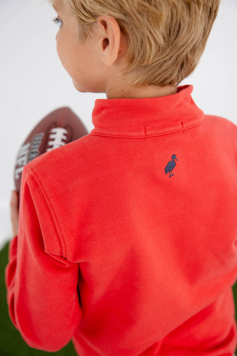 A child with blond hair, seen from behind, wears The Beaufort Bonnet Company’s Ole Miss Hayword Half-Zip in Ole Miss Red with Nantucket Navy—featuring an embroidered blue bird logo on the back—and holds an American football, making it the perfect game day layering piece.