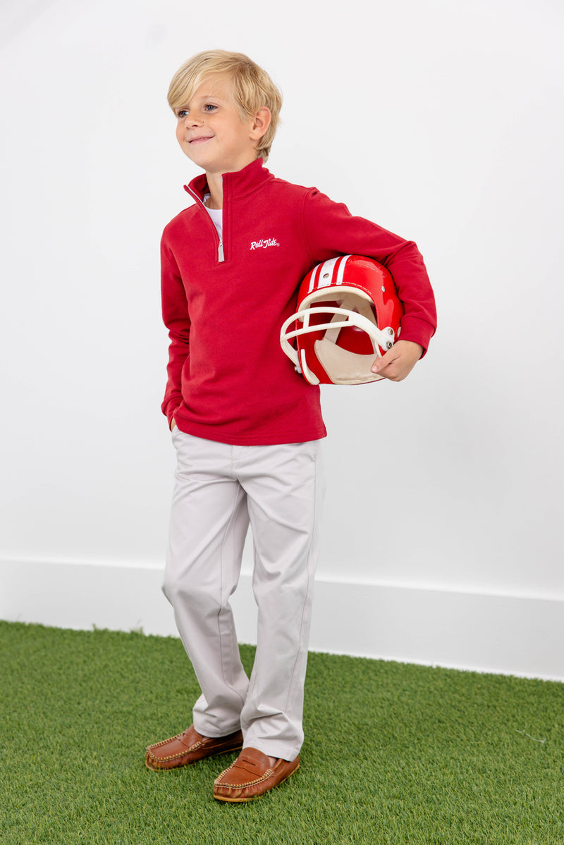Young boy in The Beaufort Bonnet Company’s Alabama Hayword Half-Zip—Alabama Crimson with Alabama Grey and Worth Avenue White—stands on green turf holding a red and white football helmet, smiling slightly, with a white wall in the background—the perfect game day layering to show off his Roll Tide spirit.