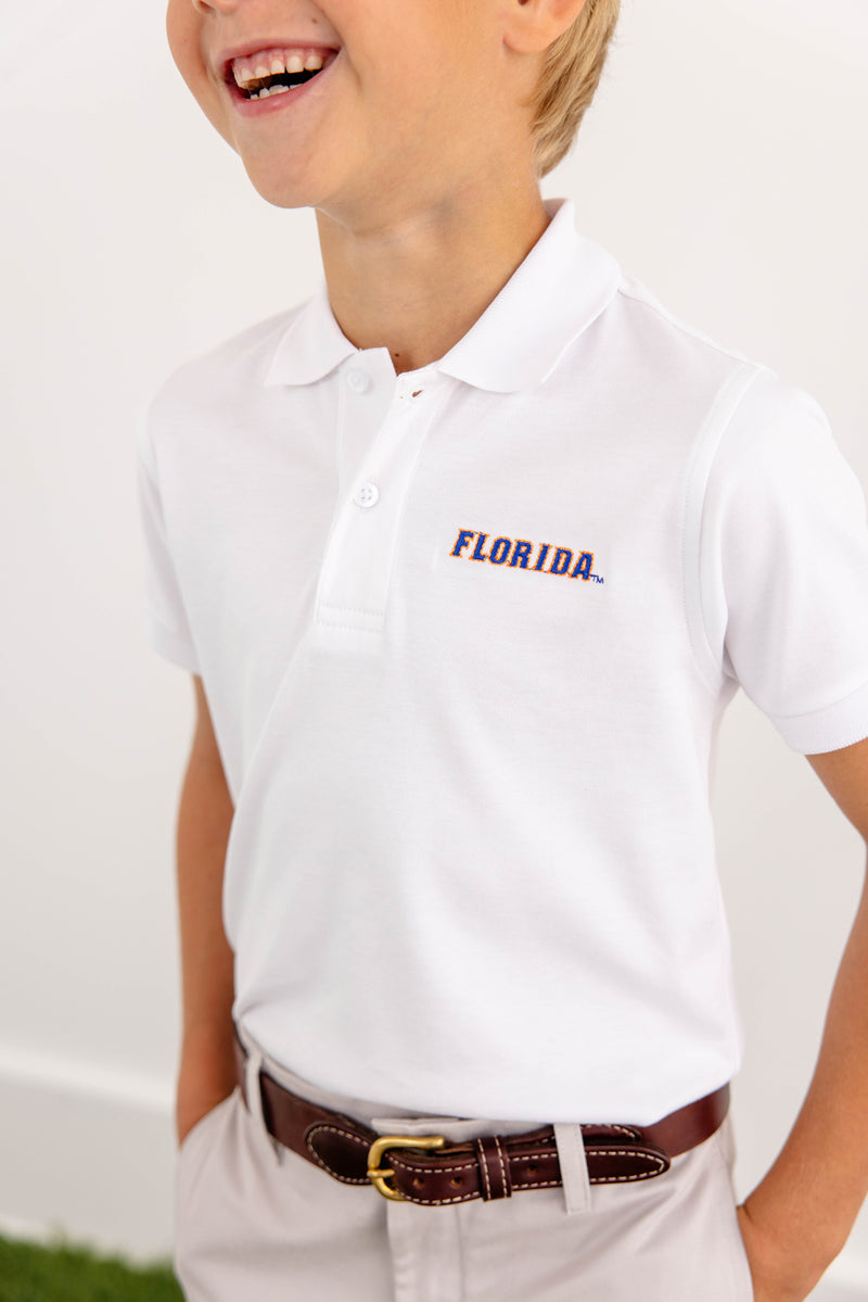 A smiling child wears The Beaufort Bonnet Company’s Florida Prim & Proper Polo in Worth Avenue White—crafted from soft Pima Cotton Knit and detailed with an embroidered Florida logo—paired with beige pants and a brown belt, set against a simple, light-colored background.