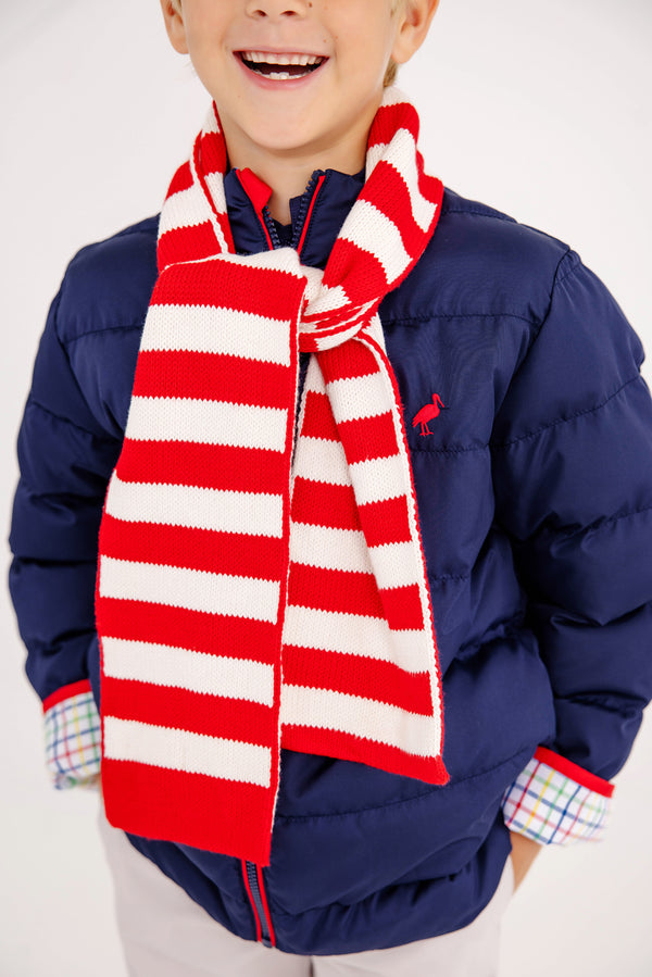 A smiling child in a blue puffer jacket with plaid shirt cuffs is wearing The Beaufort Bonnet Company’s Seller’s Scarf in Richmond Red & Palmetto Pearl Stripe.