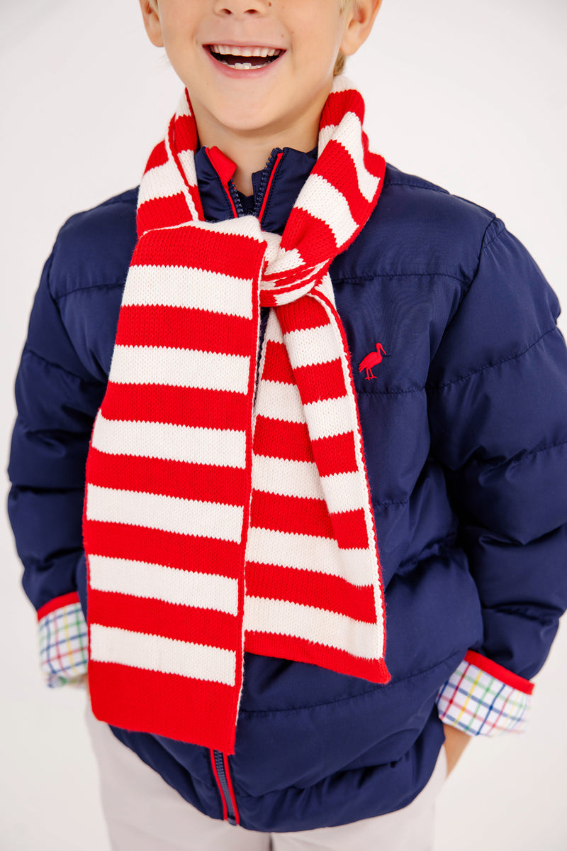 A smiling child in a blue puffer jacket with plaid shirt cuffs is wearing The Beaufort Bonnet Company’s Seller’s Scarf in Richmond Red & Palmetto Pearl Stripe.