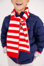 A smiling child in a blue puffer jacket with plaid shirt cuffs is wearing The Beaufort Bonnet Company’s Seller’s Scarf in Richmond Red & Palmetto Pearl Stripe.