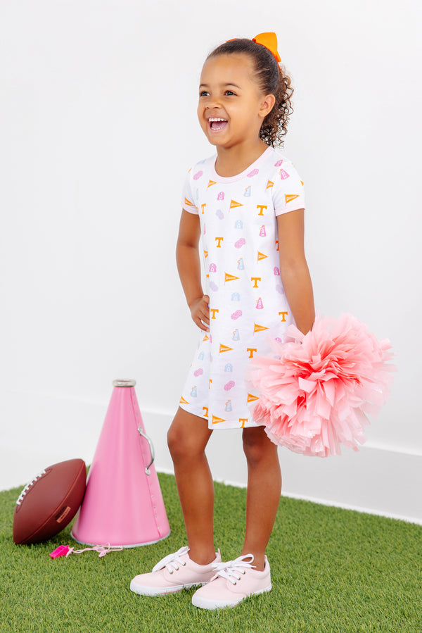 A young girl wearing the Tennessee Polly Play Dress - Victory in Orange with Palm Beach Pink from The Beaufort Bonnet Company, featuring cheerful patterns on soft Pima Cotton Knit, stands smiling while holding a large pink pom-pom. Beside her on the grass, against a white wall, lie a pink megaphone and a football.