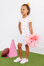 A young girl wearing the Tennessee Polly Play Dress - Victory in Orange with Palm Beach Pink from The Beaufort Bonnet Company, featuring cheerful patterns on soft Pima Cotton Knit, stands smiling while holding a large pink pom-pom. Beside her on the grass, against a white wall, lie a pink megaphone and a football.