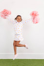 A smiling young girl jumps in the air holding pink pom-poms, wearing The Beaufort Bonnet Company’s Tennessee Searcy Skort - Victory in Orange with Worth Avenue White, paired with a white top featuring colorful patterns and pink sneakers. She stands on green artificial grass against a plain white background.