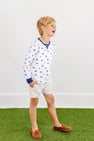 A young boy with blond hair, dressed in a white patterned long-sleeve shirt, khaki shorts, and brown loafers, stands on green artificial grass and smiles while looking to the side against a plain white background. He completes his look with The Beaufort Bonnet Company’s Ole Miss Cassidy Comfy Crewneck - Gawsh Almighty with Nantucket Navy for a fun and casual style.