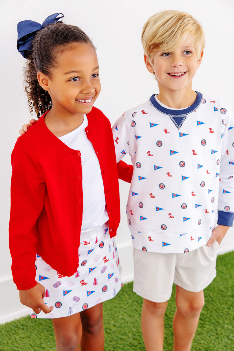 Two children stand side by side, smiling. The girl wears a red cardigan, white shirt, patterned skirt, and a blue bow in her hair. The boy wears The Beaufort Bonnet Company’s Ole Miss Cassidy Comfy Crewneck - Gawsh Almighty with Nantucket Navy and light shorts. They stand on green grass indoors.