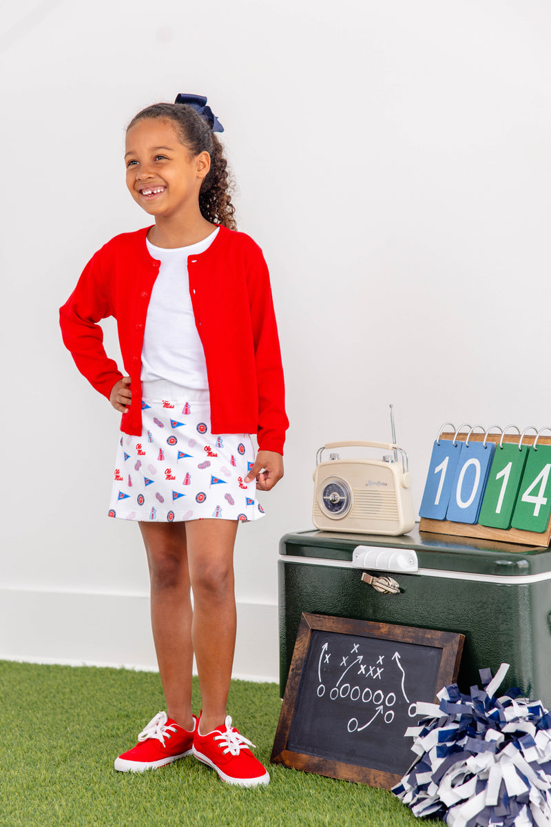 A smiling girl wearing a red cardigan, white shirt, and The Beaufort Bonnet Company’s Ole Miss Searcy Skort – Gawsh Almighty with Worth Avenue White stands on artificial grass. Beside her are a chalkboard displaying football plays, a radio, scorecards, and pom-poms for a sports-themed scene.